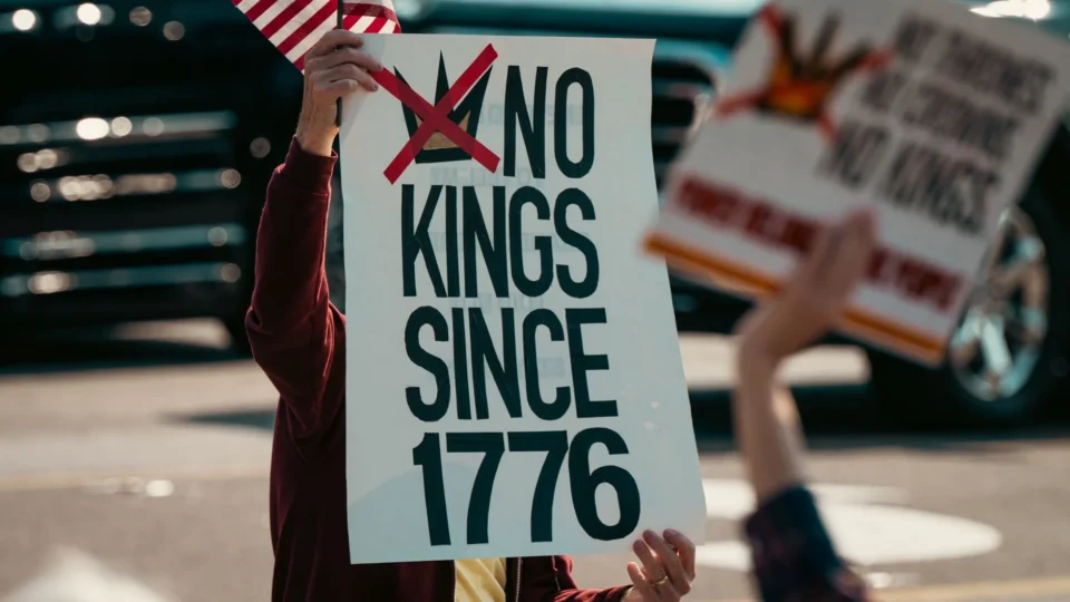 Protest sign displaying 'No Kings Since 1776' held alongside a small US flag, emphasizing democratic sentiments.