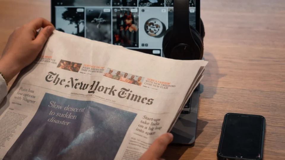 Close-up of hands holding a newspaper by a laptop and smartphone on a table.