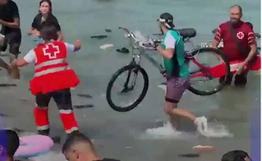 A migrant carrying a bicycle through shallow water as Red Cross workers assist nearby