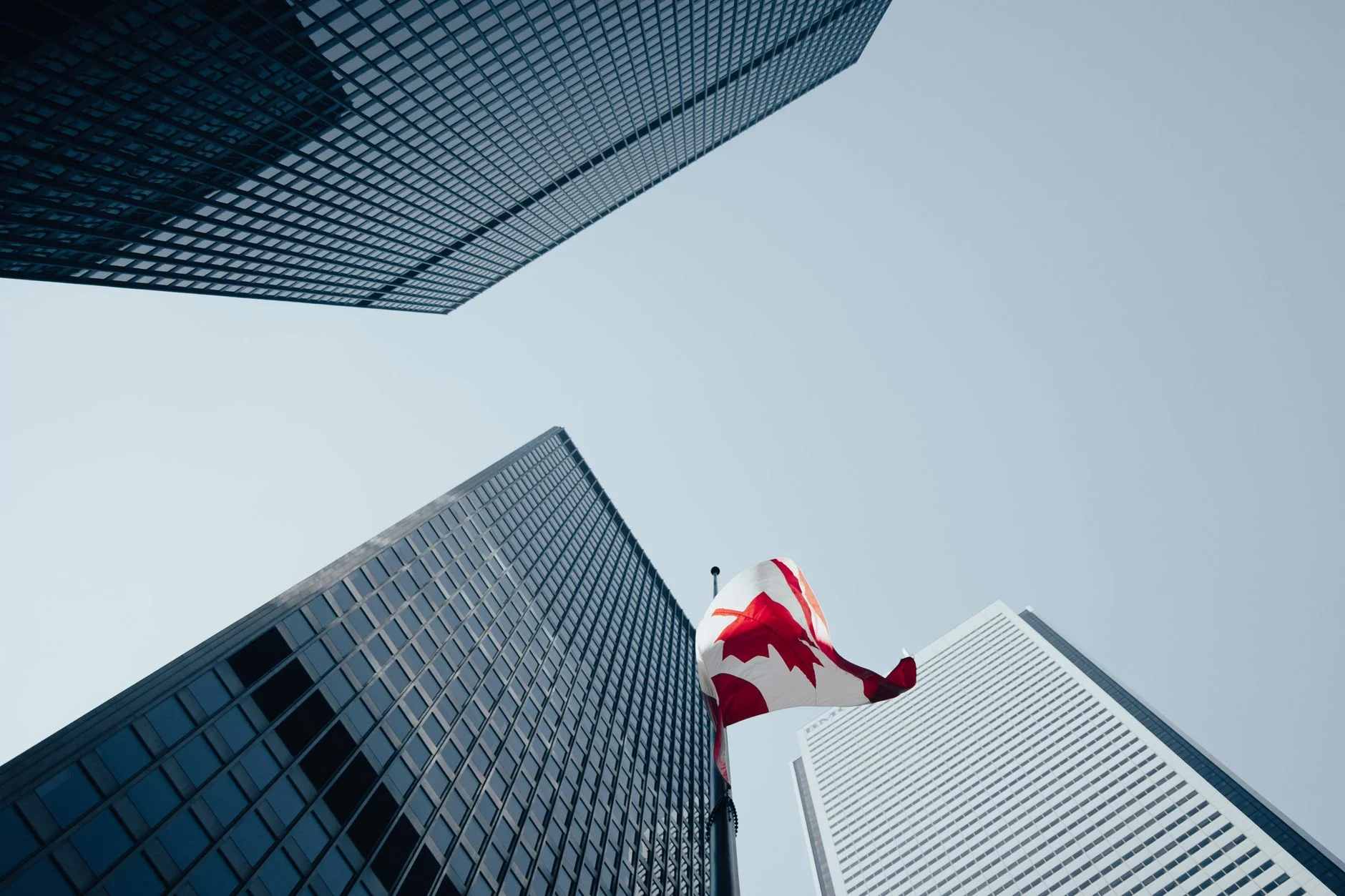 Dramatic low-angle view of skyscrapers and a Canadian flag in downtown Toronto, under a clear blue sky.