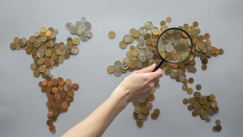 Top view of crop unrecognizable traveler with magnifying glass standing over world map made of various coins on gray background