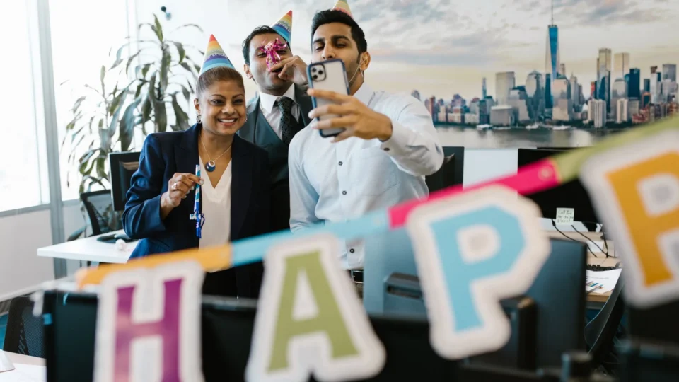 Three colleagues celebrating a birthday in the office with party hats and decorations, capturing a fun selfie.