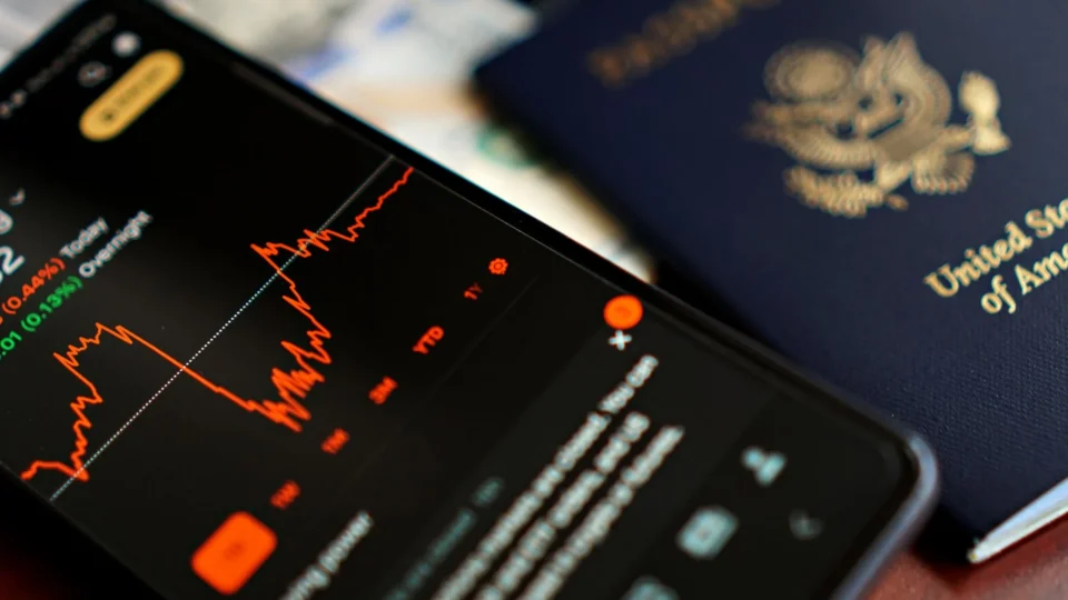 Close-up of a smartphone displaying stock data next to a US passport, suggesting travel and finance themes.