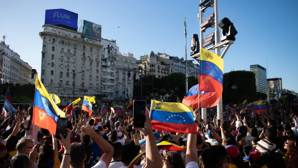 A large crowd waves Venezuelan flags during a protest in a bustling city square.