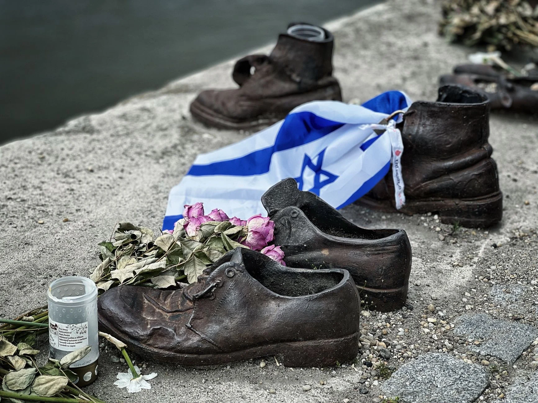 Sculpture of shoes with Israeli flag and flowers on riverbank, symbolizing remembrance.