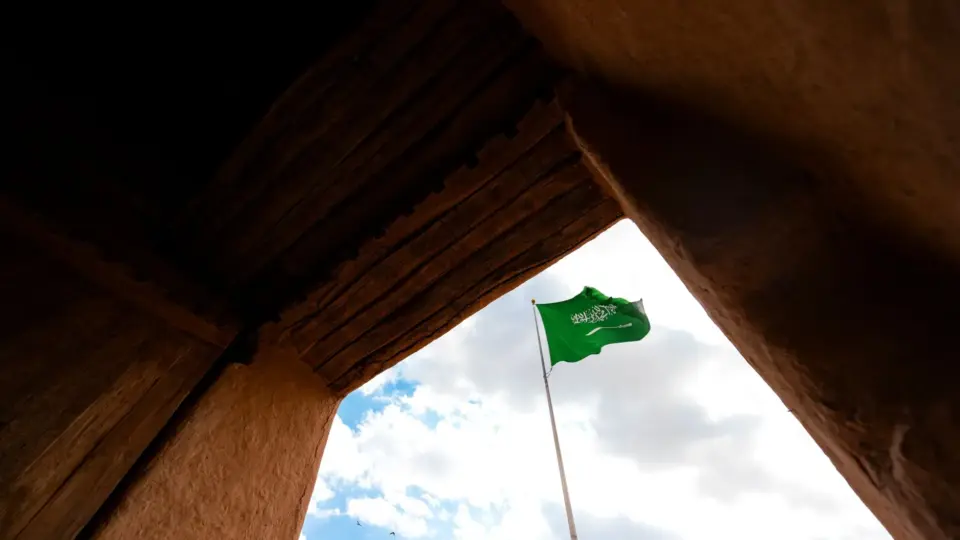 Saudi Arabian flag waves against blue sky amid traditional architecture.